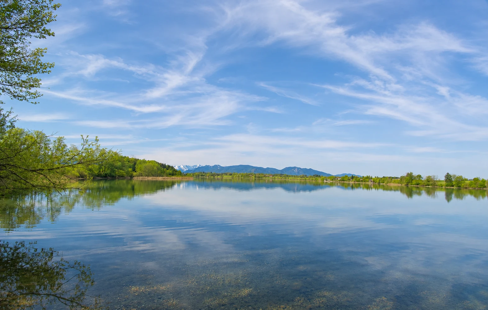 Ruhiger See mit Spiegelungen, grünen Ufern und Bergen im Hintergrund.