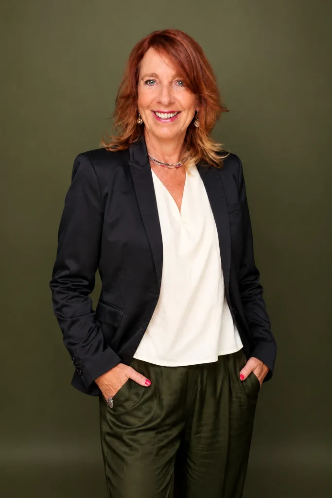 Ilona Gotz im schwarzen Blazer und weißer Bluse, mit den Händen in den Hosentaschen, vor grünem Hintergrund.